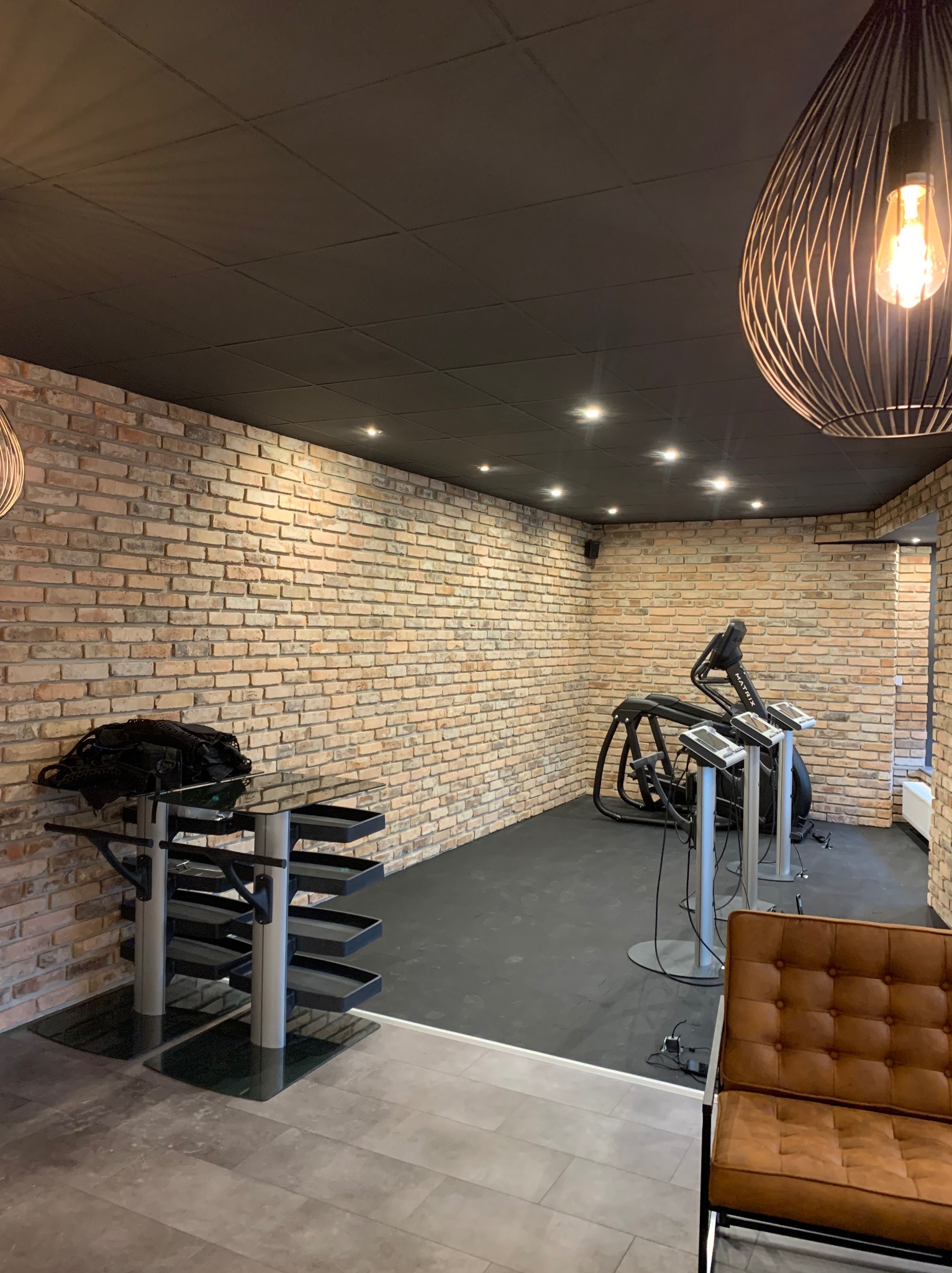 Wnętrze pomieszczenia z ceglaną ścianą, czarnym sufitem kasetonowym z oświetleniem punktowym, metalową lampą wiszącą z żarówką Edisona, brązową sofą i sprzętem do ćwiczeń.
