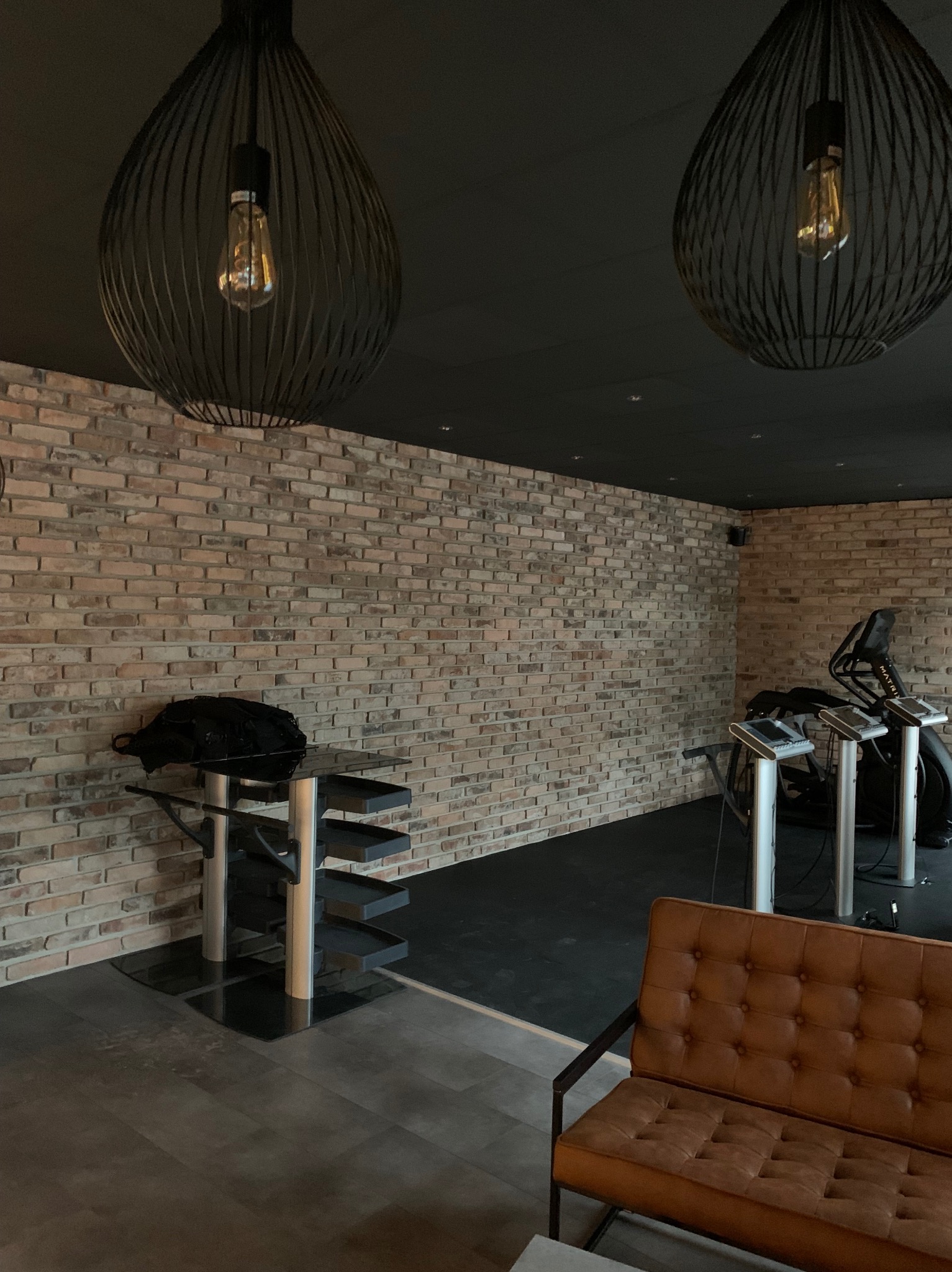 Surowe ceglane ściany z dekoracyjnymi lampami wiszącymi, brązowa sofa i sprzęt do ćwiczeń w tle, tworzące nowoczesną przestrzeń.