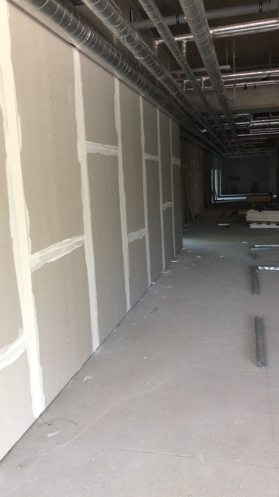 Montaż płyt gipsowo-kartonowych na ścianie w przestrzeni komercyjnej, widoczne szpachlowane łączenia i instalacja wentylacyjna pod sufitem.