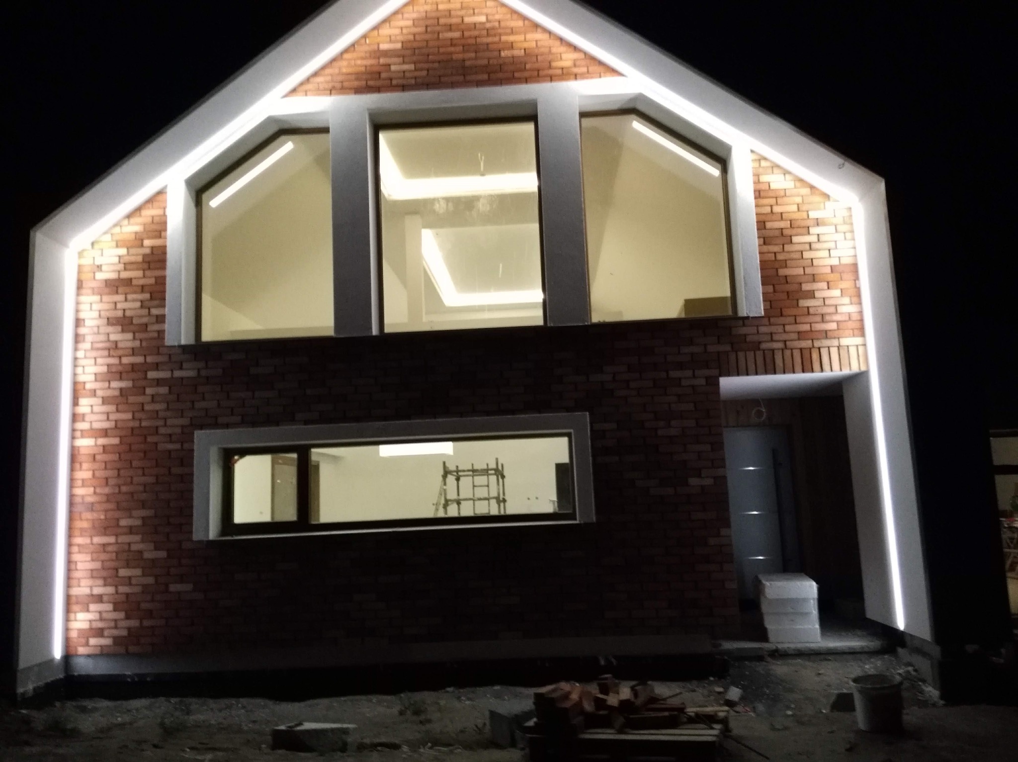 Dwukondygnacyjny budynek z cegły z oknami oświetlonymi od wewnątrz i na zewnątrz białym światłem LED, widoczne rusztowanie wewnątrz budynku przez okno na parterze.