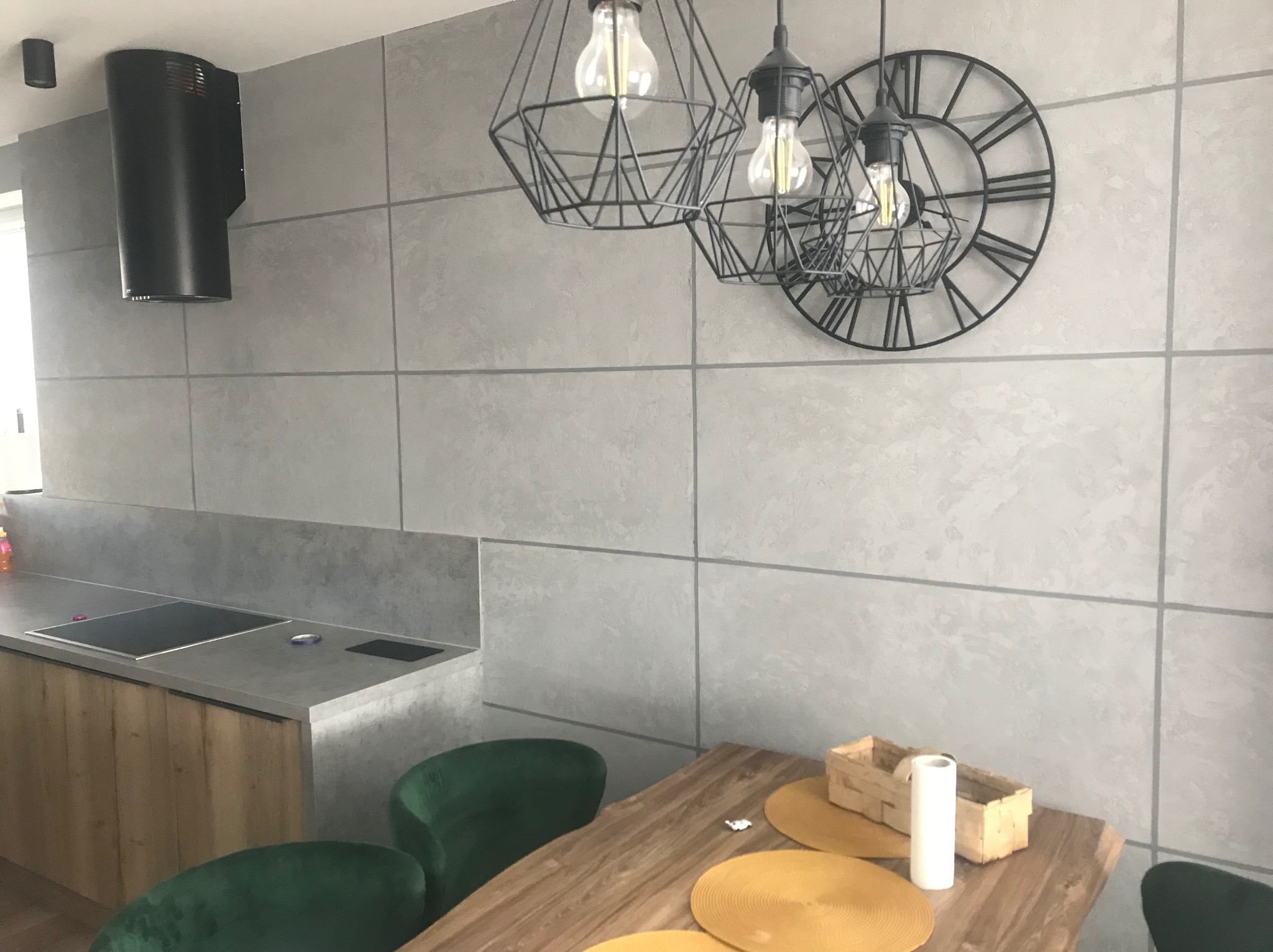 Szara kuchnia z wyspą, zielonymi krzesłami, geometrycznymi lampami i zegarem na ścianie z szarych płyt.