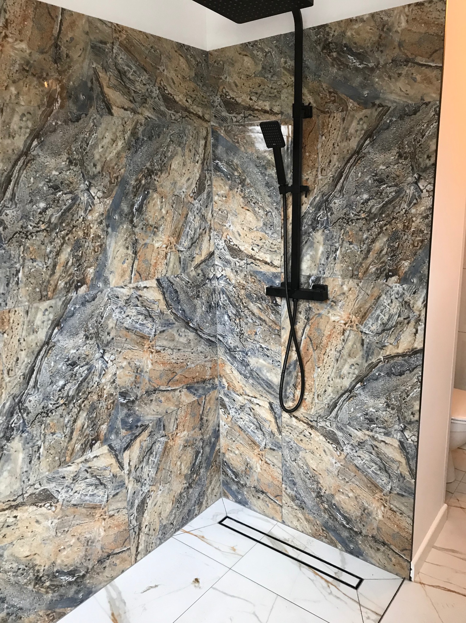 Kabina prysznicowa z czarną armaturą i płytkami imitującymi marmur w odcieniach szarości, beżu i brązu, widoczny odpływ liniowy w podłodze wyłożonej białymi płytkami z szarymi żyłkami.