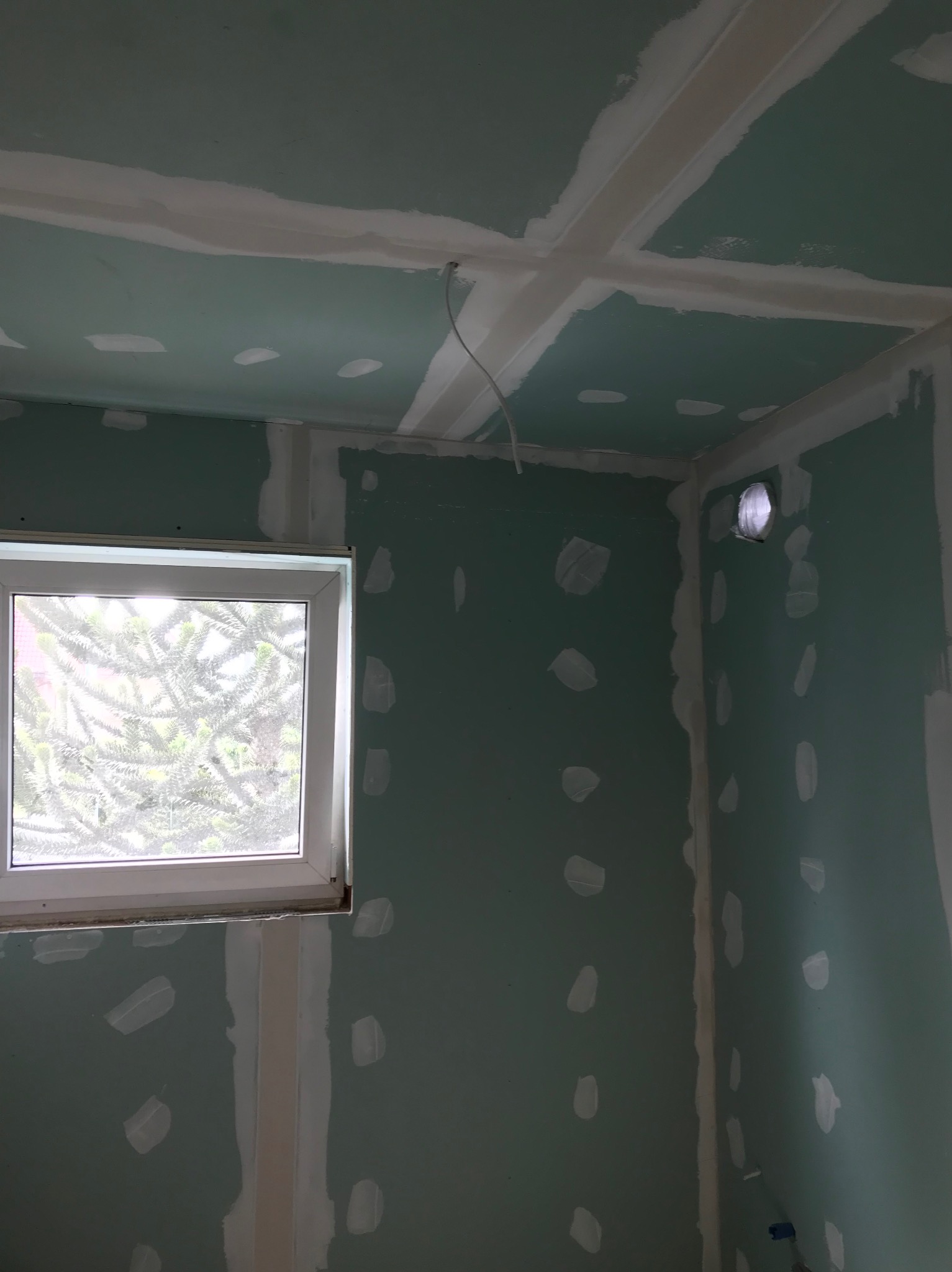 Ściany i sufit z płyt gipsowo-kartonowych w trakcie szpachlowania, widoczne łączenia płyt, otwór wentylacyjny w rogu, okno z widokiem na zieleń.