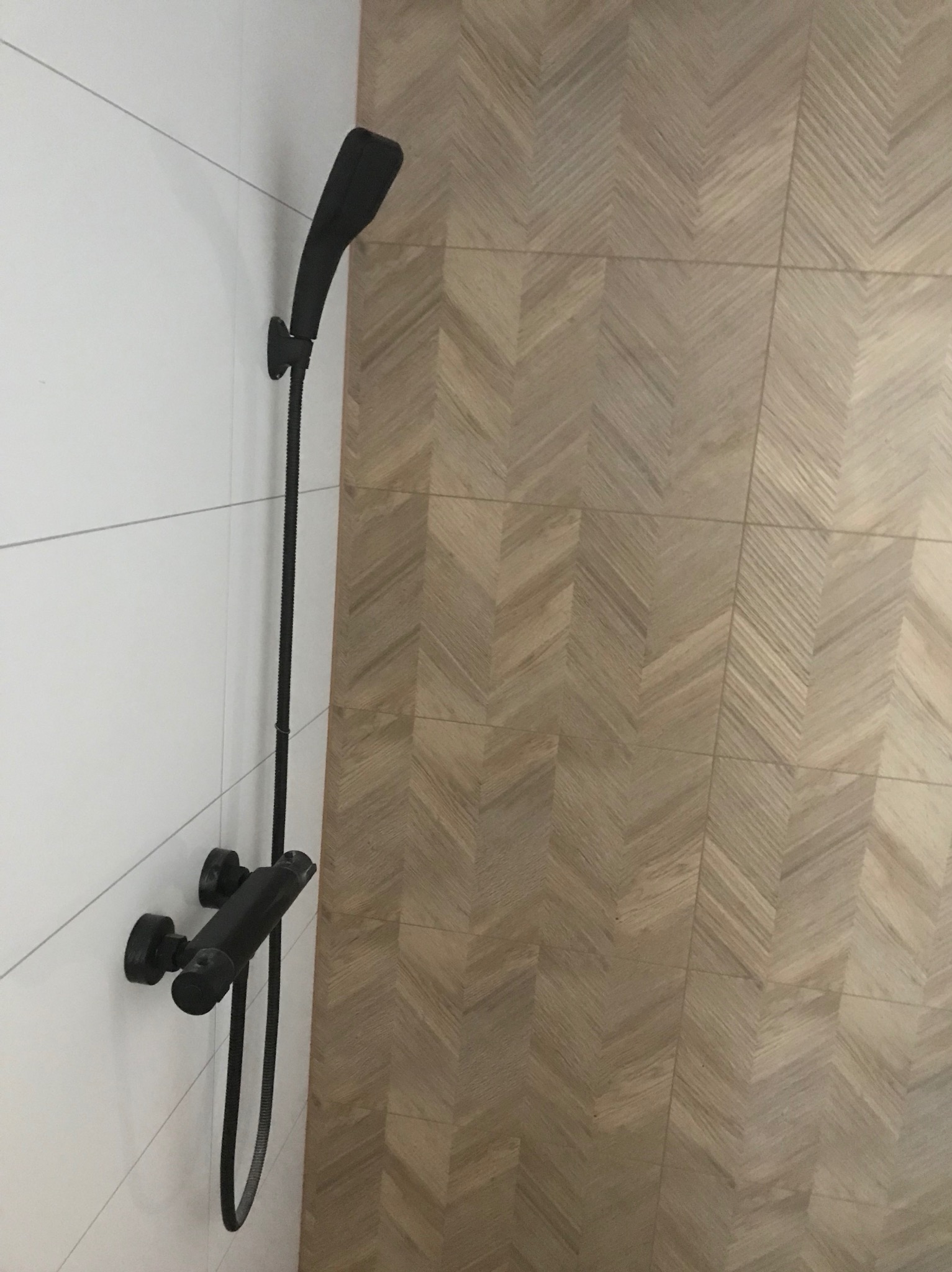 Czarny zestaw prysznicowy zamontowany na białej, kafelkowanej ścianie, z podłogą wyłożoną parkietem w jodełkę w tle.