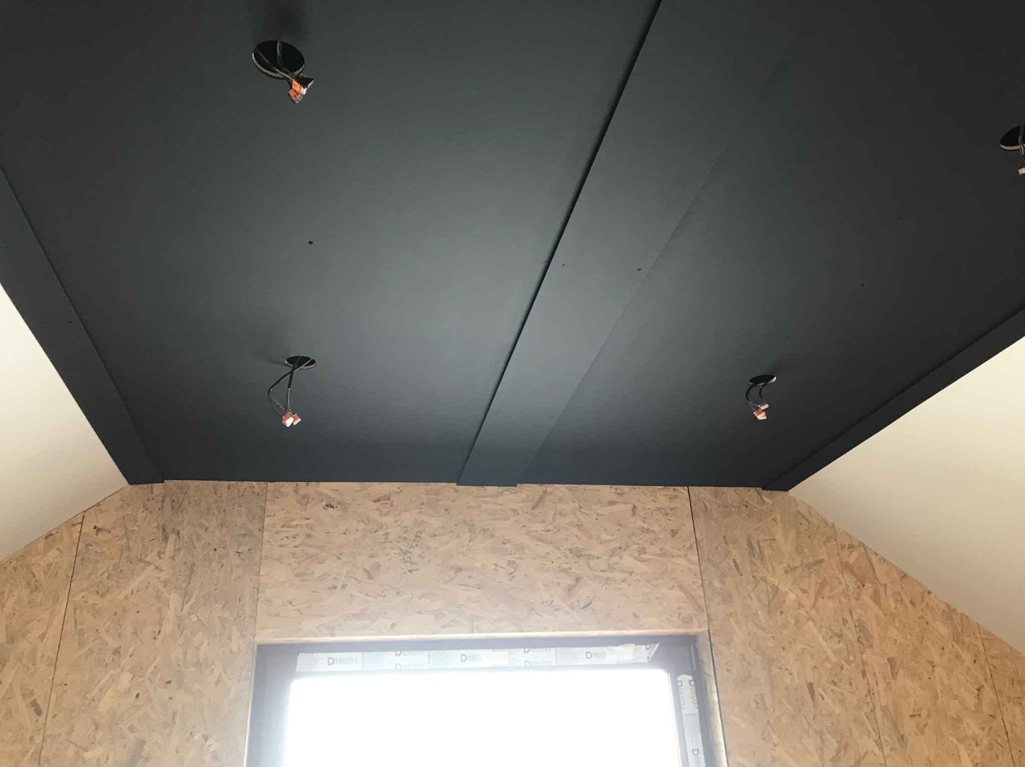 Ciemny, matowy sufit z widocznymi przewodami oświetleniowymi w surowym wnętrzu poddasza z oknem i ścianami wyłożonymi płytą OSB.