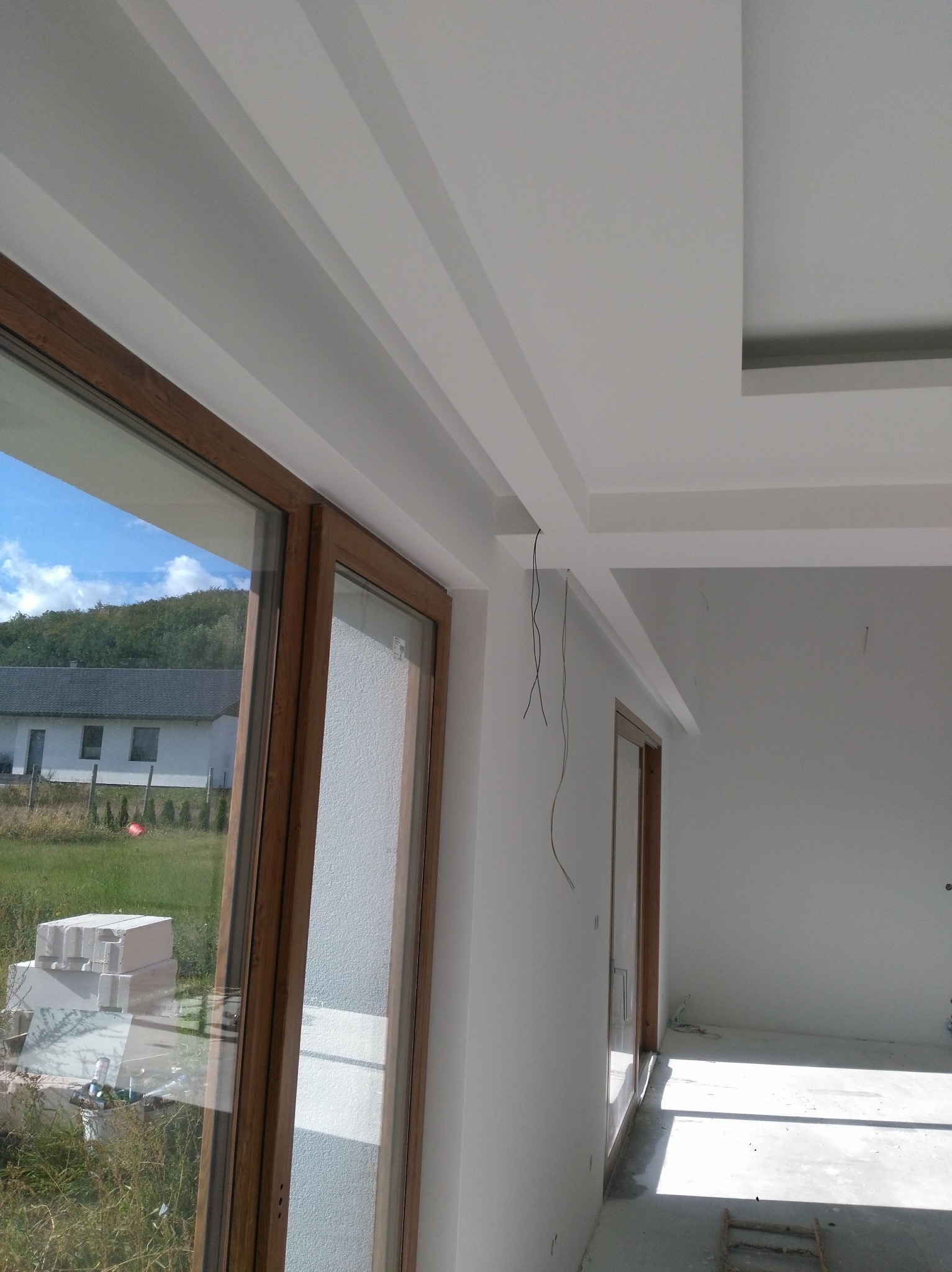 Nowoczesny pokój w trakcie wykańczania: białe ściany, drewniane okna z widokiem na zewnątrz, dekoracyjny sufit podwieszany z oświetleniem, widoczne kable i materiały budowlane na zewnątrz.