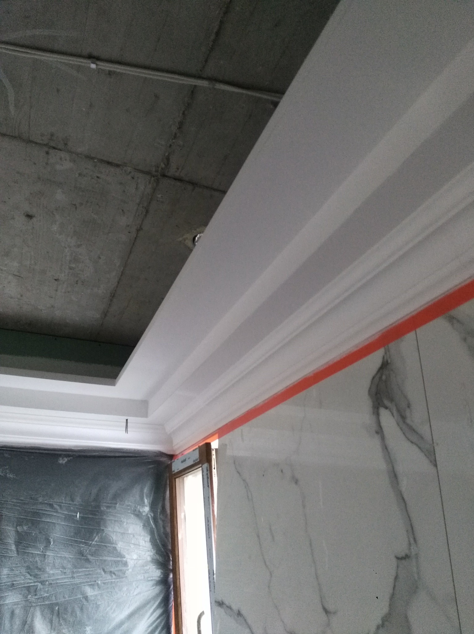 Montaż ozdobnych listew przysufitowych w pomieszczeniu z surowym betonowym stropem i marmurowymi płytkami ściennymi, zabezpieczonym folią oknem.