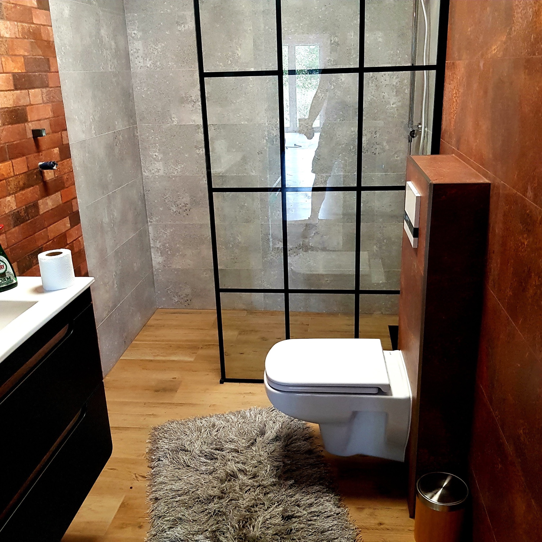Nowoczesna łazienka z kabiną prysznicową typu walk-in z czarnymi szprosami, wisząca toaleta, drewnopodobna podłoga, ściana z cegły i szarych płytek oraz brązowa ściana z ukrytą spłuczką.