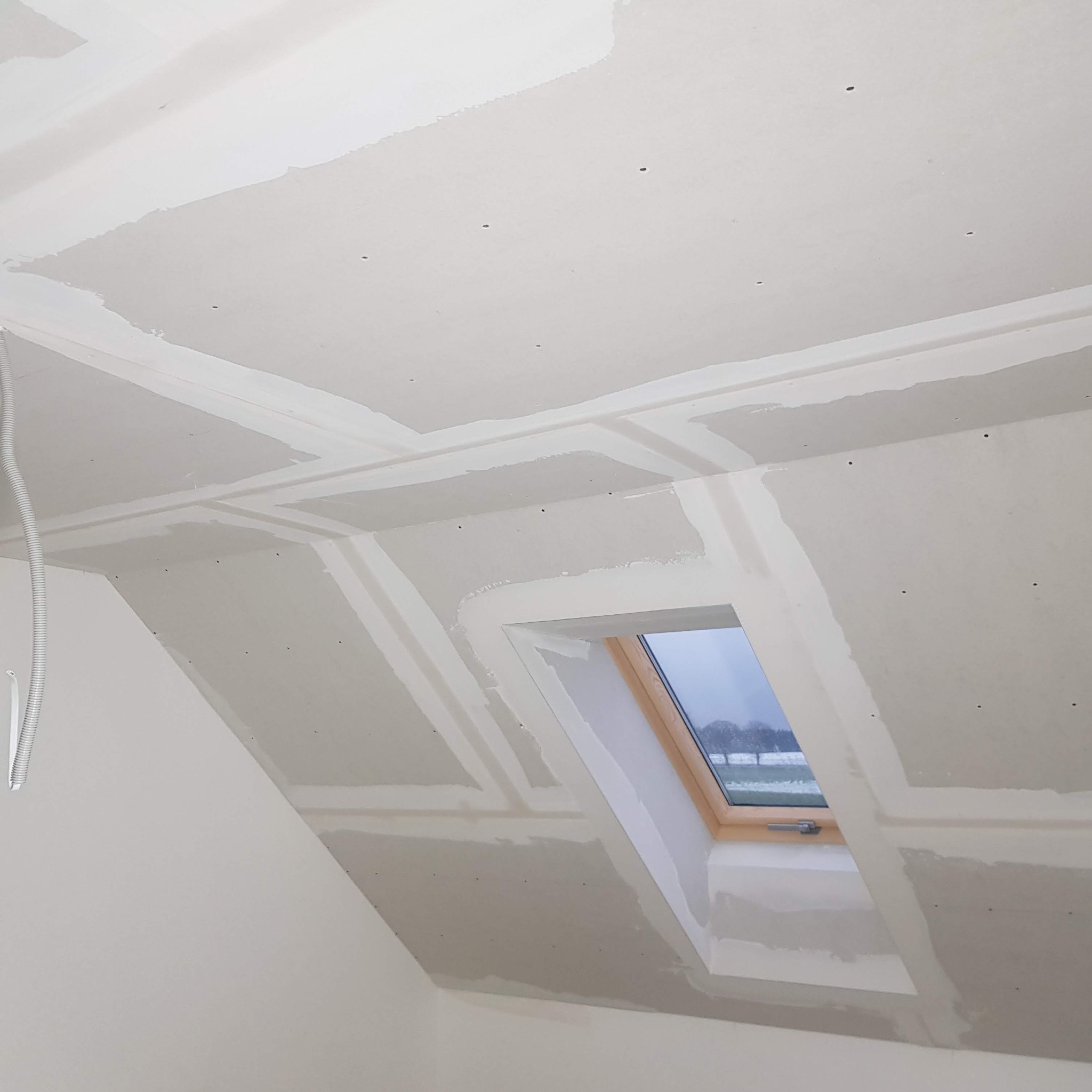 Poddasze w Pucku w trakcie wykańczania: widoczne płyty gipsowo-kartonowe ze szpachlowanymi łączeniami oraz okno dachowe z widokiem na zaśnieżone pole.