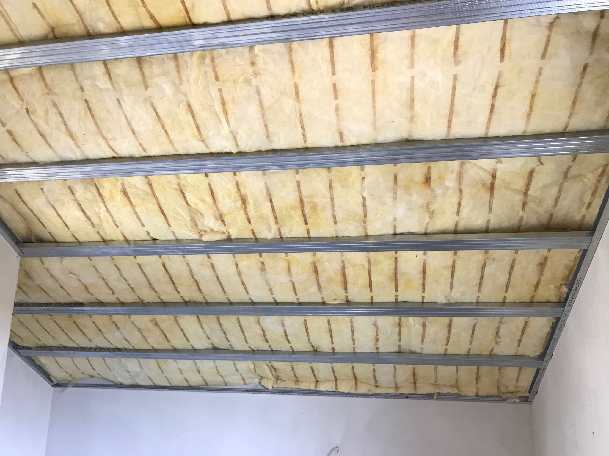 Poddasze w trakcie ocieplania: wełna mineralna umieszczona między metalowymi profilami konstrukcji sufitu podwieszanego, widoczne niedociągnięcia w ułożeniu izolacji.