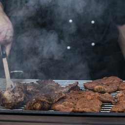 Catering JW Olejniczak - Kucharz w czarnym uniformie obraca szczypcami mięso na grillu, unoszący się dym i soczyste steki w zbliżeniu.