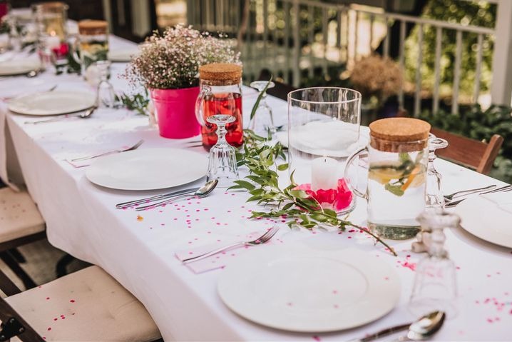 Uroczysty stół z białym obrusem, nakryty porcelanową zastawą, srebrnymi sztućcami, szklankami na nóżce, dzbankami z korkami i napojami, różowym wiaderkiem z gipsówką oraz konfetti w kształcie serc...