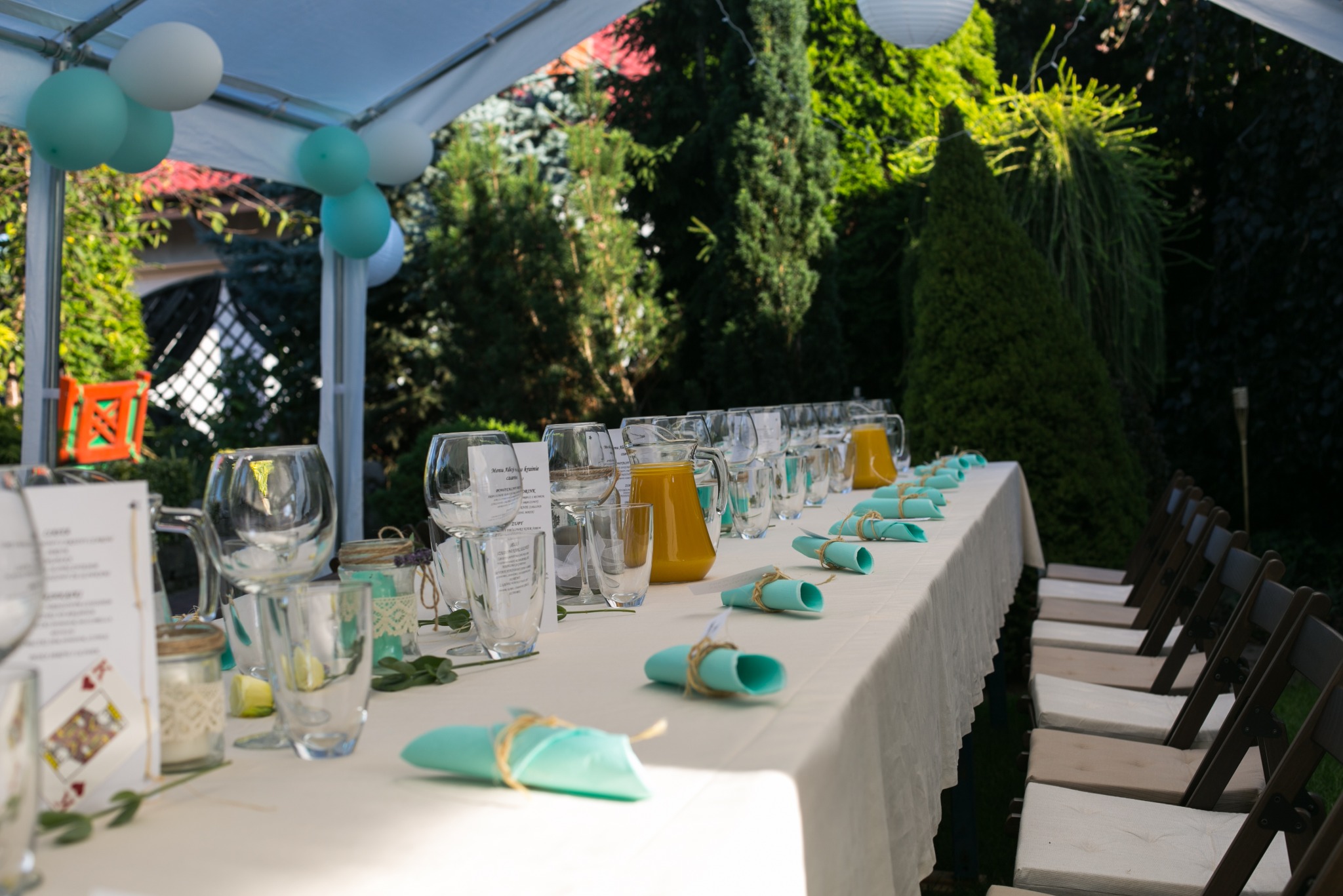 Uroczysty stół pod namiotem, nakryty białym obrusem, z ustawionymi szklankami, dzbankami z sokiem pomarańczowym i turkusowymi serwetkami związanymi sznurkiem, otoczony zielenią ogrodu i dekoracjami...