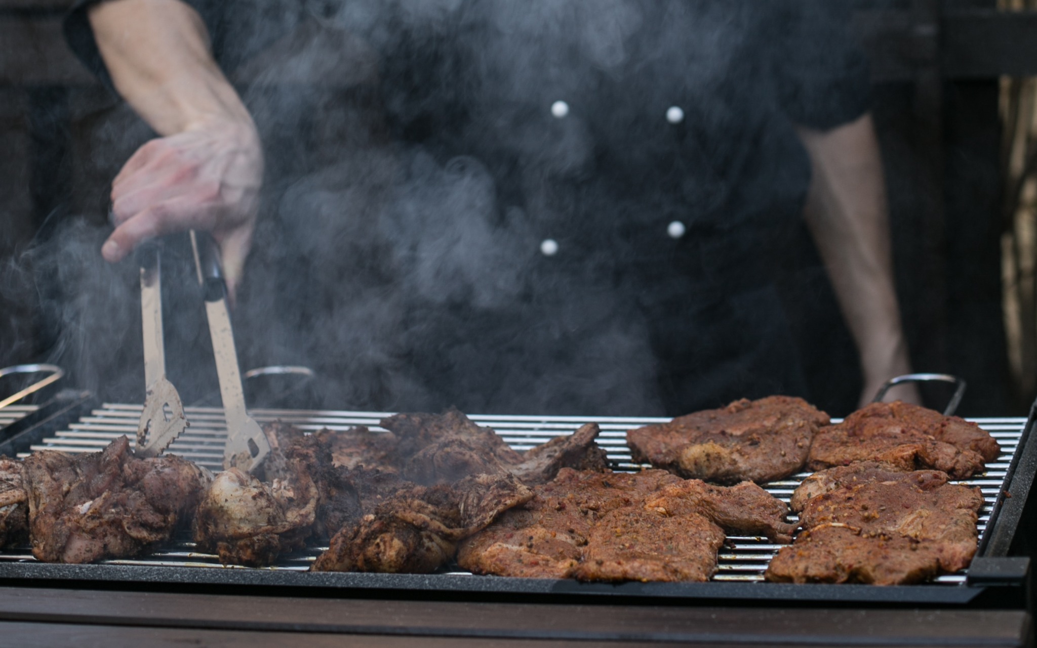 Kucharz w czarnym uniformie obraca szczypcami mięso na grillu, unoszący się dym i soczyste steki w zbliżeniu.