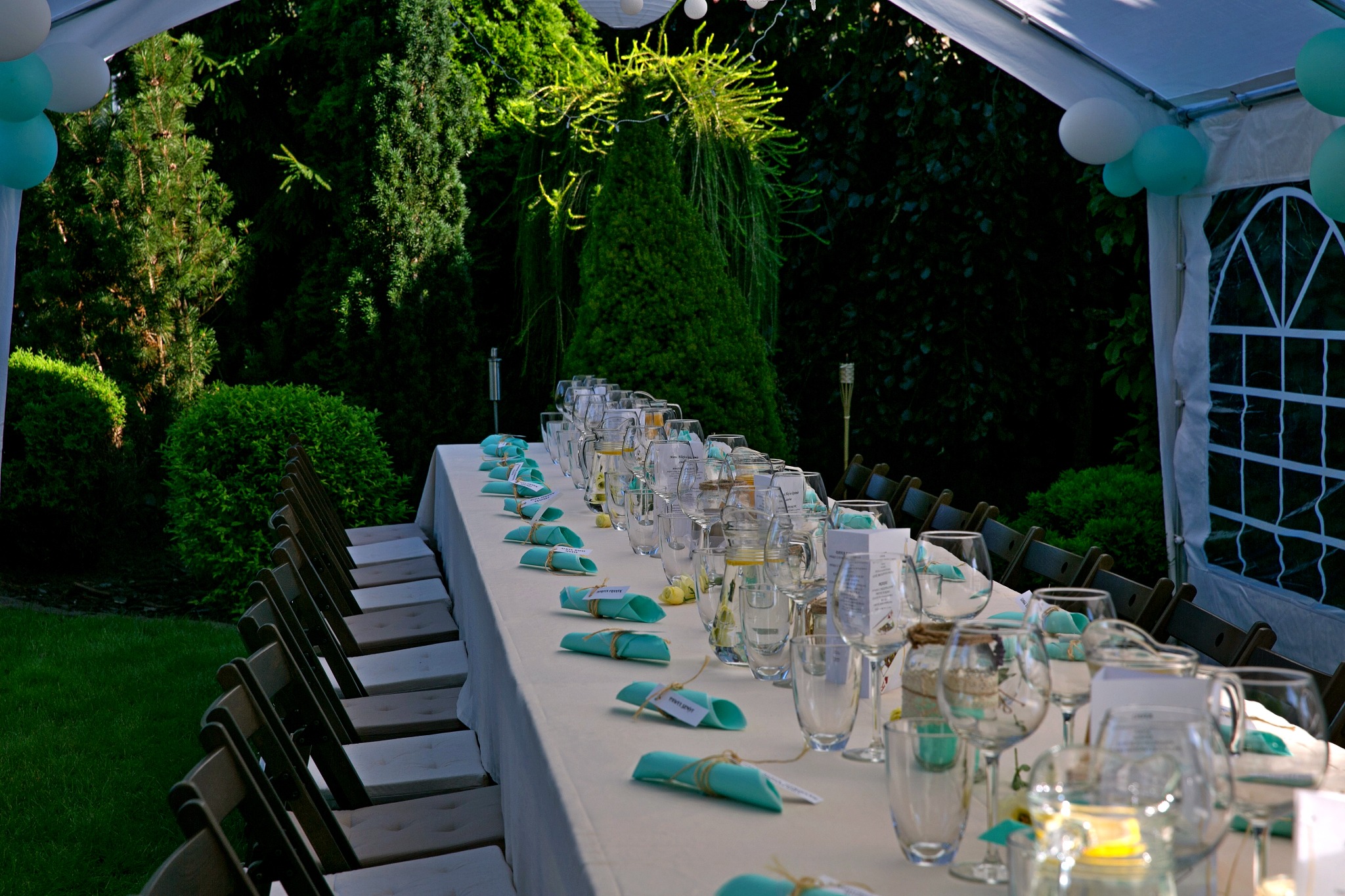 Długi stół pod białym namiotem, nakryty białym obrusem, z turkusowymi serwetkami i szklanymi naczyniami, otoczony zielenią ogrodu, przygotowany na przyjęcie.