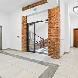 ReJoN Nieruchomości Joanna Rejnowicz - Jasne wnętrze budynku w Gliwicach z windą, schodami widocznymi przez otwarte drzwi i korytarzem z drewnianymi drzwiami, wykończone białymi ścianami i jasną podłogą z szarymi akcentami.
