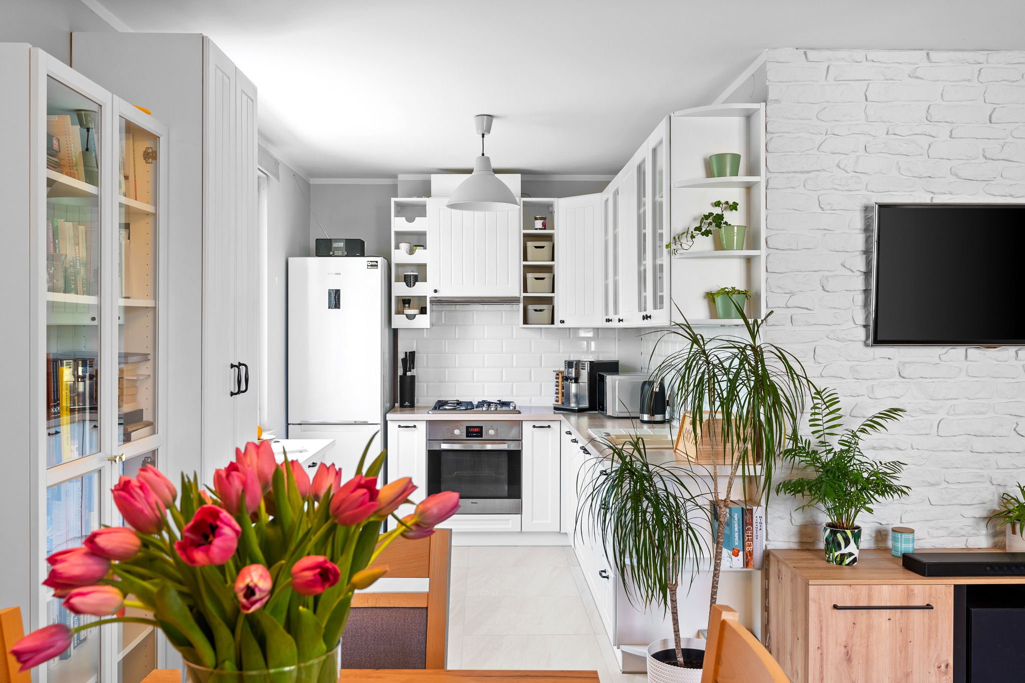 Jasna kuchnia z białymi meblami, ceglaną ścianą i bukietem różowych tulipanów na pierwszym planie, widok na sprzęty AGD i dekoracyjne rośliny.
