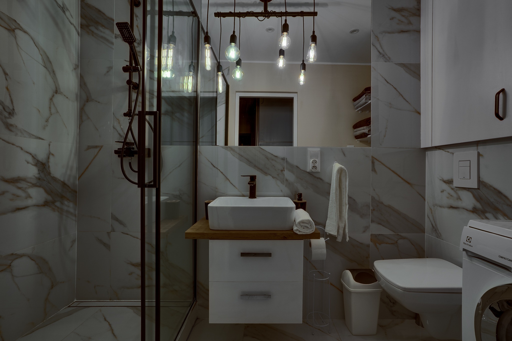 Nowoczesna łazienka z marmurowymi płytkami, kabiną prysznicową w ciemnej ramie, umywalką na drewnianym blacie, wiszącymi żarówkami Edison i pralką Electrolux.
