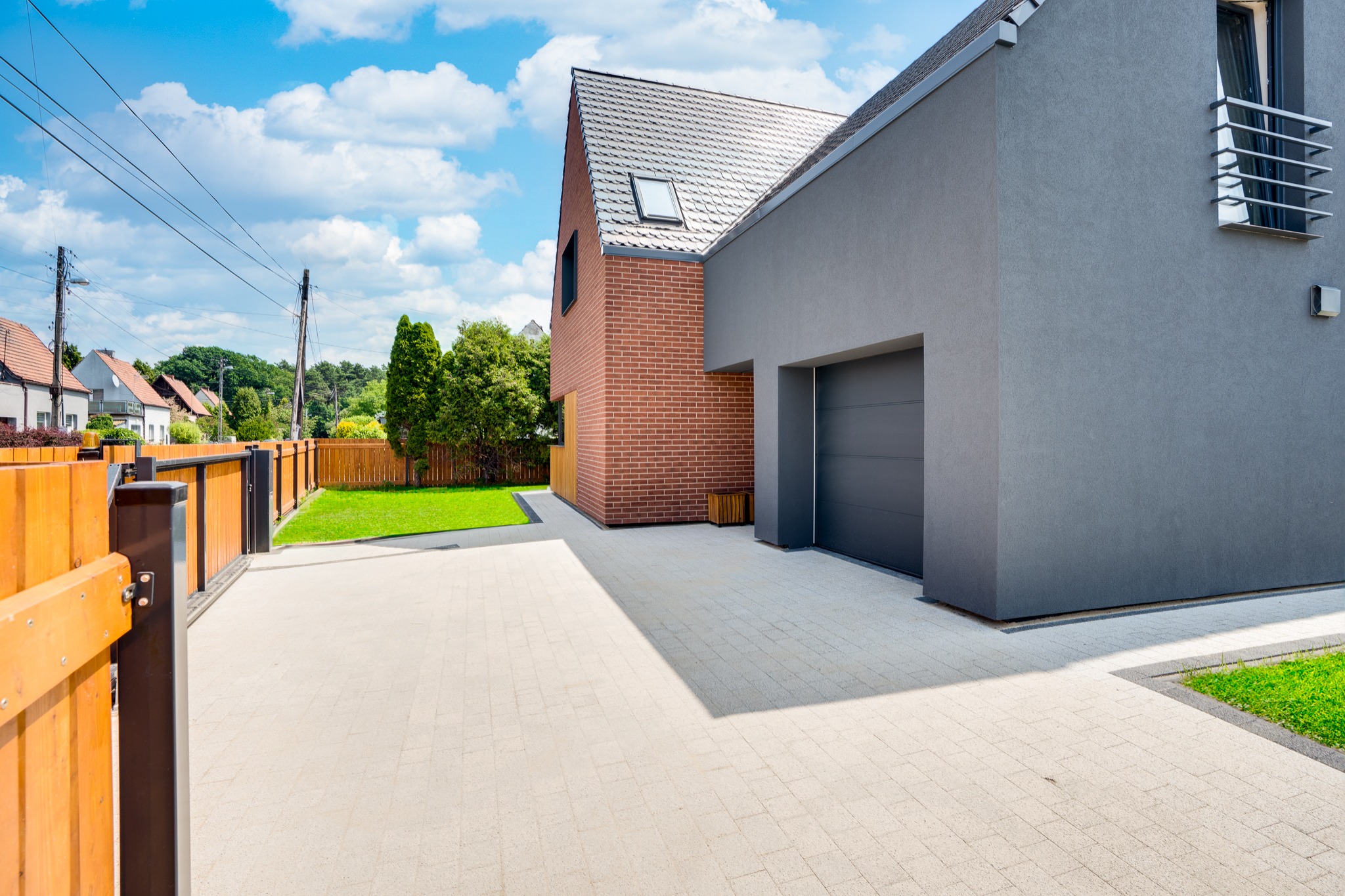 Nowoczesny dom z elewacją w kolorze szarości i cegły, z garażem, podjazdem z kostki brukowej i drewnianym ogrodzeniem w pogodny dzień.