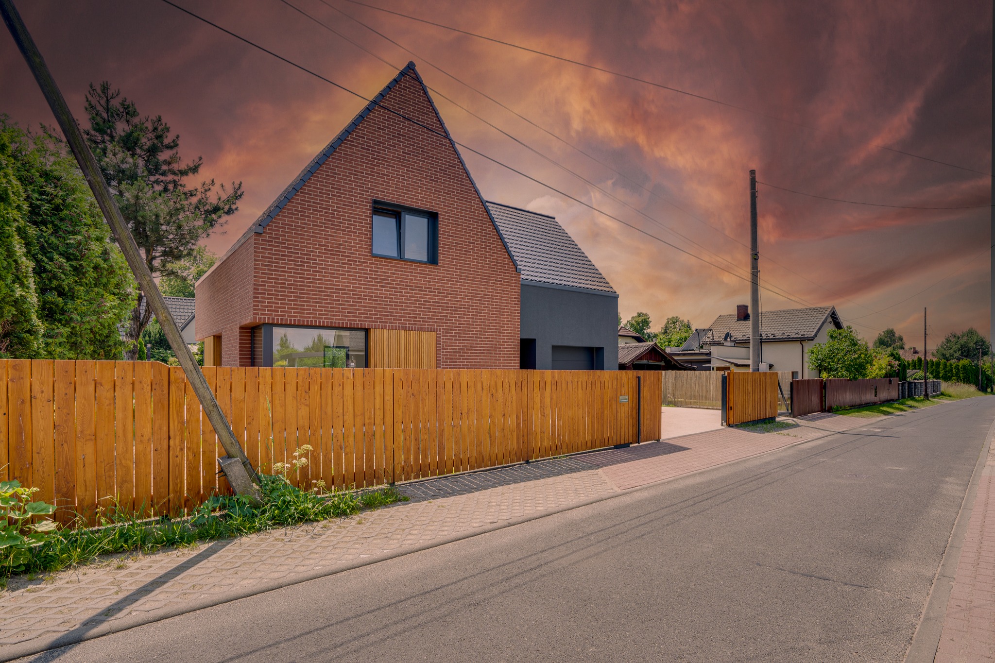 Nowoczesny dom jednorodzinny z cegły i szarego tynku, drewniane ogrodzenie, brama wjazdowa, widok z ulicy, dynamiczne chmury na niebie.