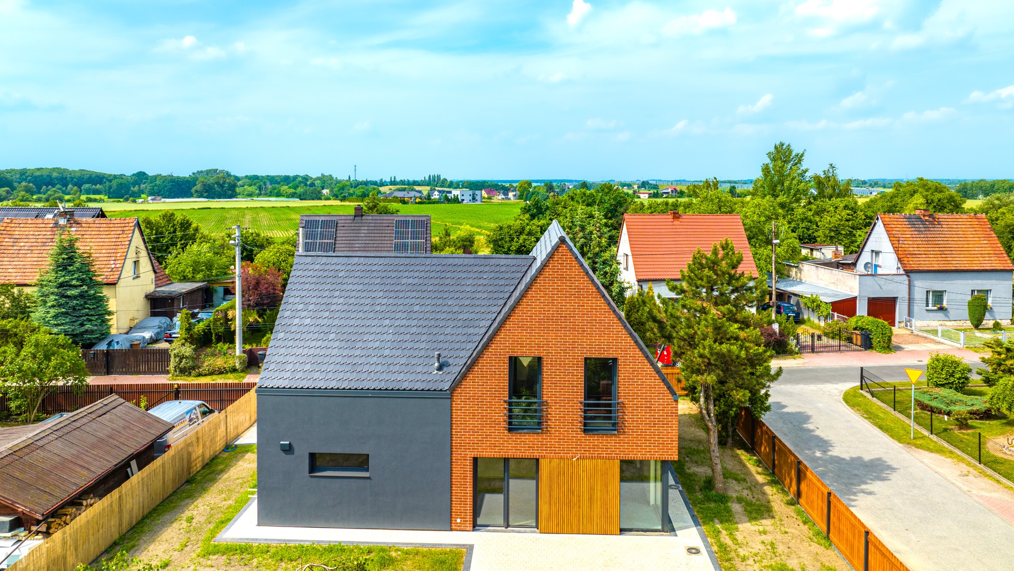 Nowoczesny dom jednorodzinny z dwuspadowym dachem, elewacją z cegły i szarego tynku, widok z góry na tle zieleni i innych zabudowań.
