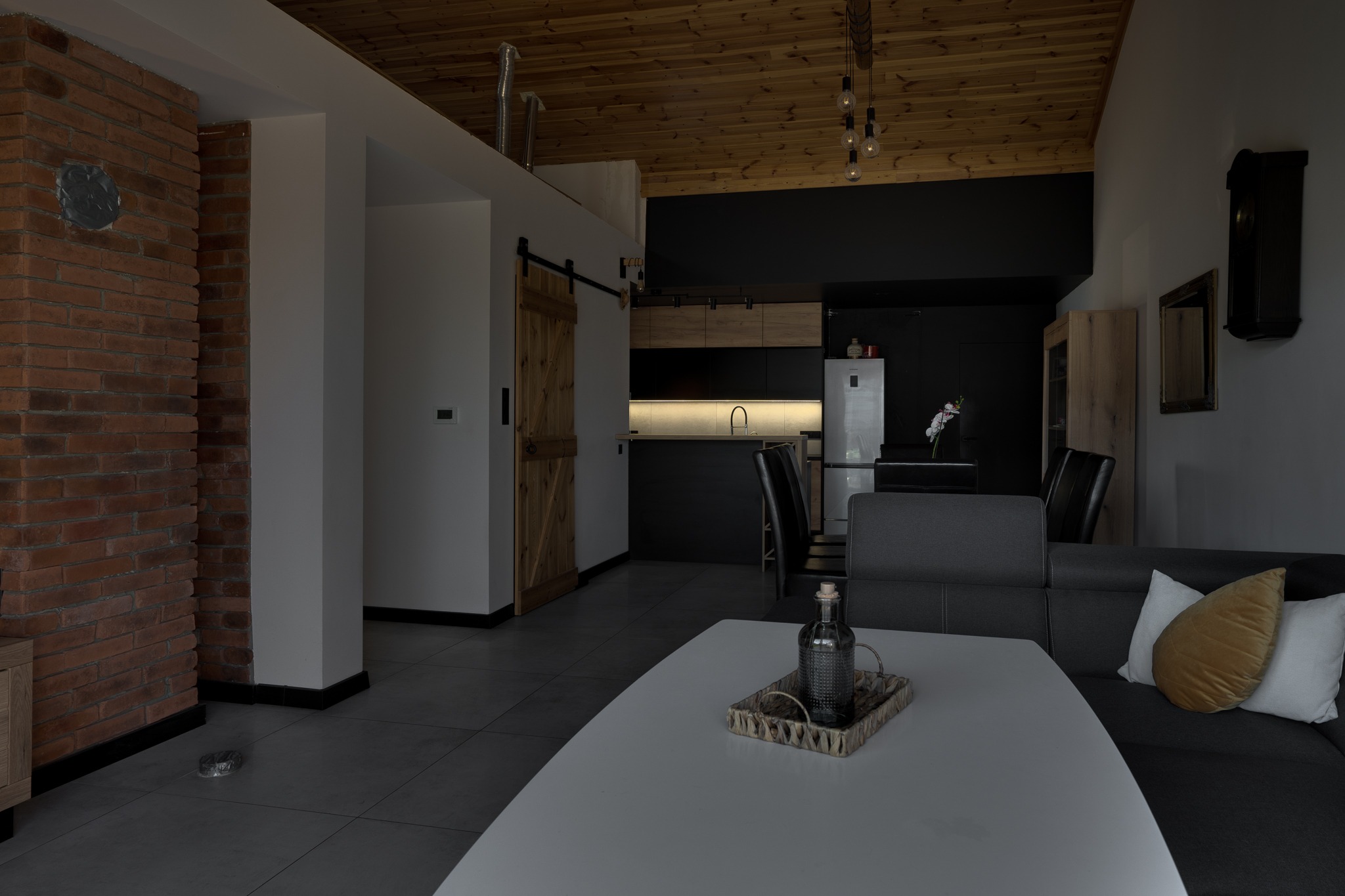 Nowoczesny salon z ceglaną ścianą, drewnianym sufitem i przesuwnymi drzwiami stodołowymi, minimalistyczna kuchnia w tle, butelka dekoracyjna na białym stoliku kawowym.