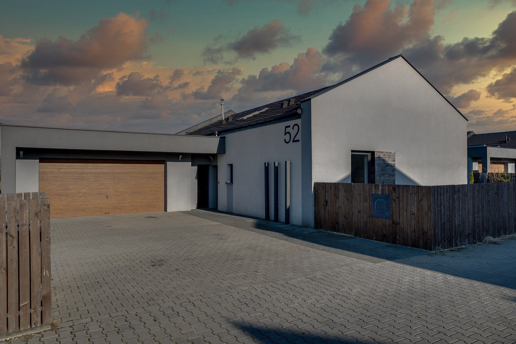 Nowoczesny dom jednorodzinny z garażem i drewnianym ogrodzeniem, numer domu 52 widoczny na elewacji, kostka brukowa na podjeździe, zachodzące słońce tworzy dramatyczne tło.