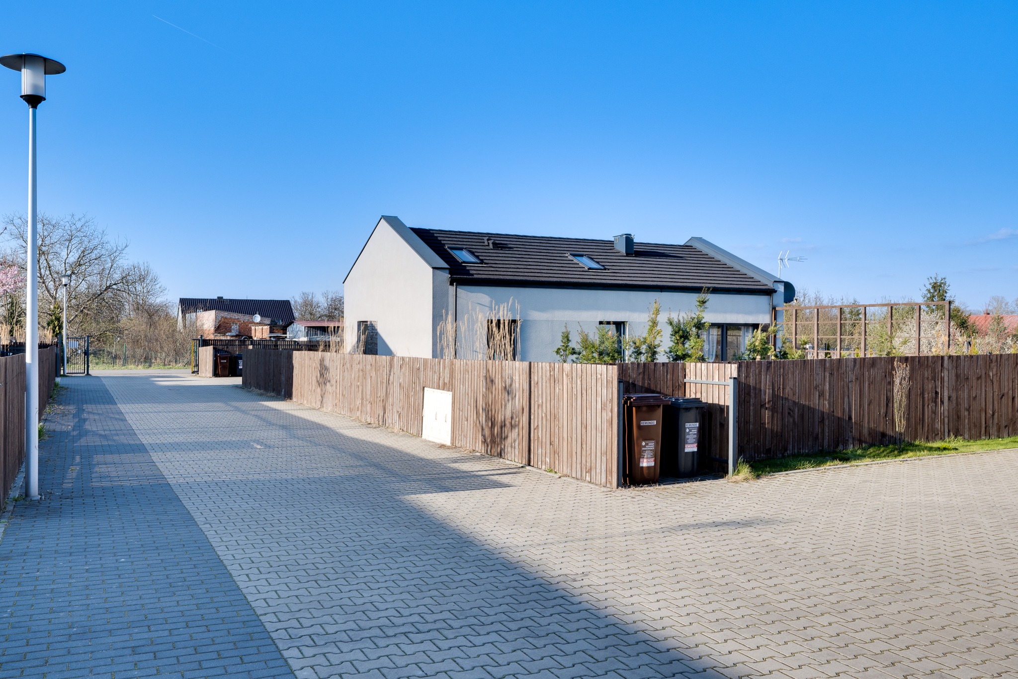 Nowoczesny dom jednorodzinny z szarą elewacją i ciemną dachówką, ogrodzony drewnianym płotem, z dwoma pojemnikami na śmieci przy wjeździe, widziany z brukowanej drogi w słoneczny dzień.