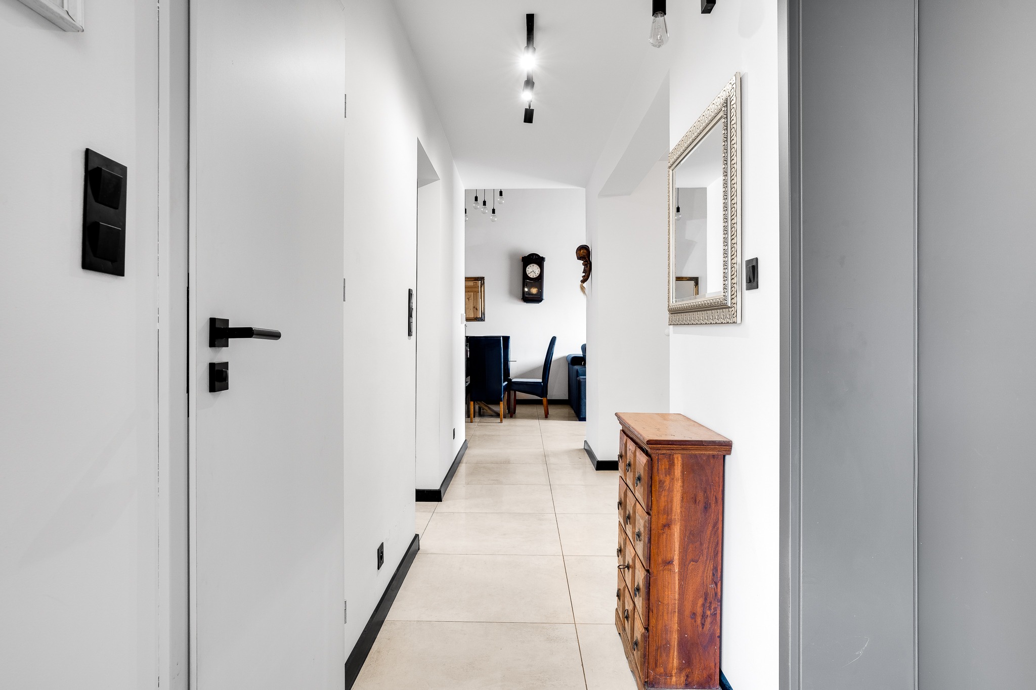 Nowoczesny korytarz z białymi ścianami, drewnianą komodą, lustrem w ozdobnej ramie, czarnymi akcentami i widokiem na salon z zegarem i lampami
