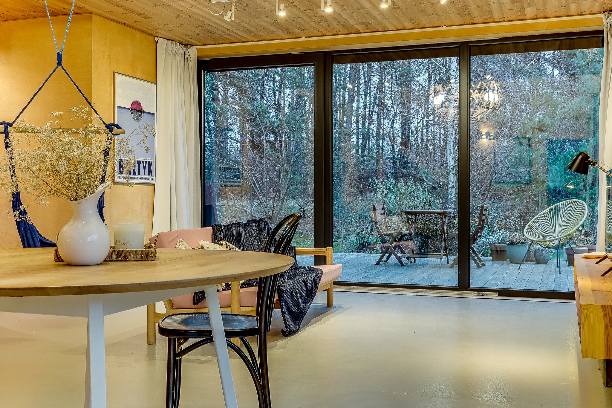 Jasne, przestronne wnętrze z drewnianym sufitem i dużym oknem tarasowym wychodzącym na ogród; okrągły stół z białym wazonem i suszonymi kwiatami w centrum, w tle hamak i minimalistyczny plakat.