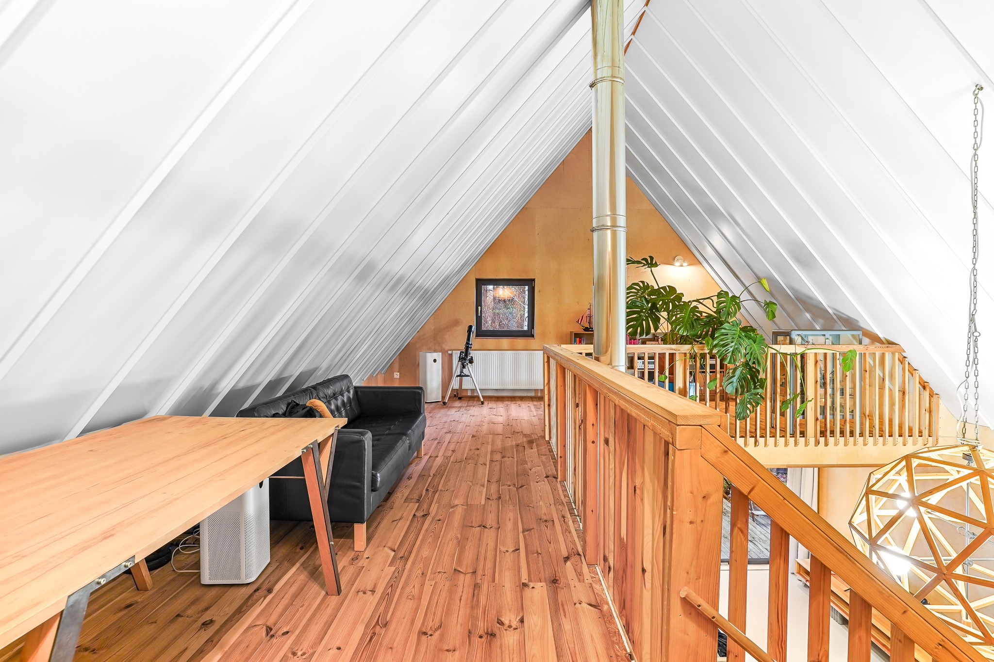 Przestronne poddasze z drewnianą podłogą, skośnym białym sufitem, czarną sofą, biurkiem z jasnego drewna i roślinami doniczkowymi, oświetlone naturalnym światłem wpadającym przez okno.