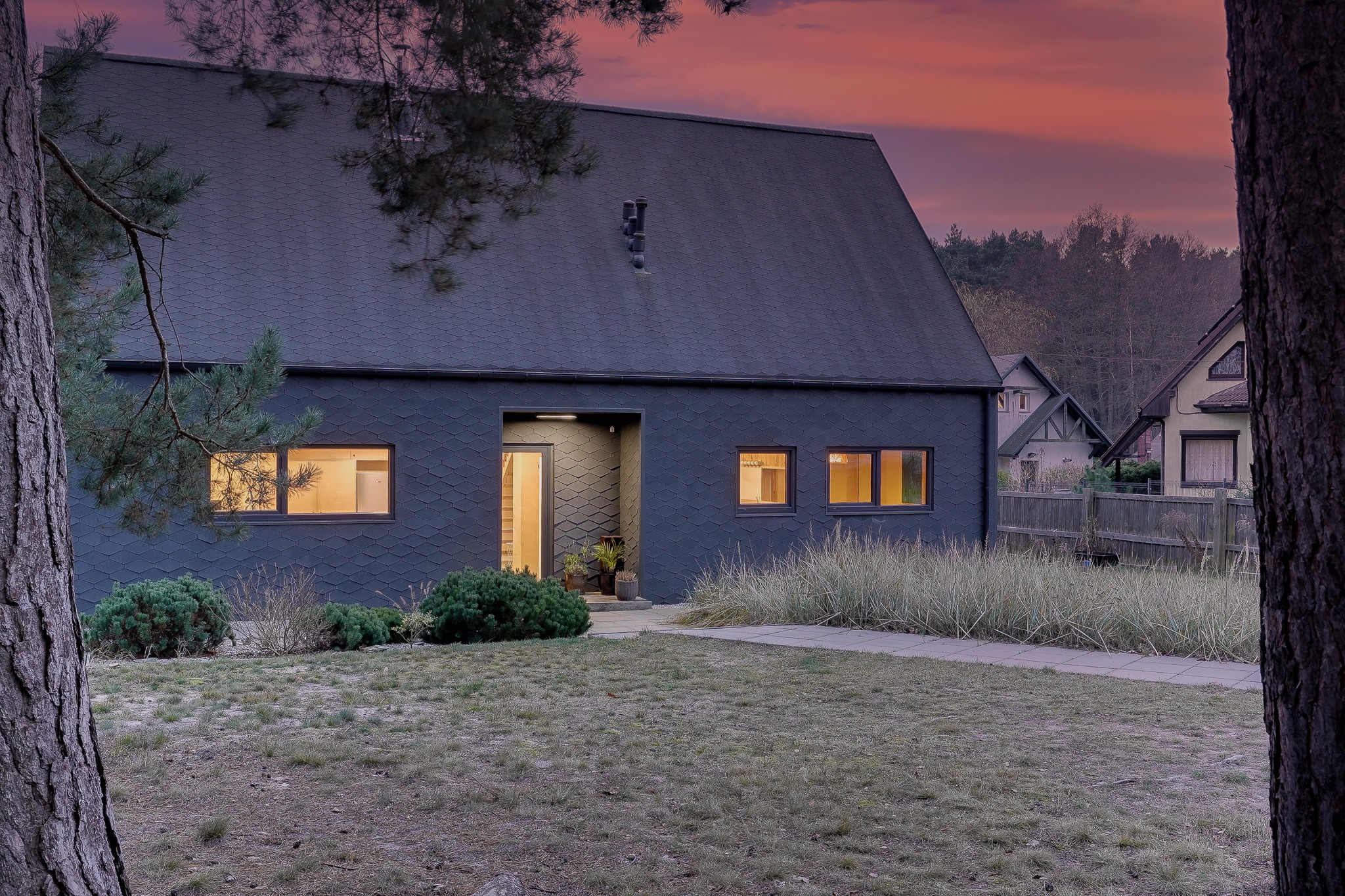 Nowoczesny dom jednorodzinny z dachem i elewacją pokrytą ciemną dachówką karpiówką, oświetlony ciepłym światłem z okien, widoczny z perspektywy ogrodu o zmierzchu.