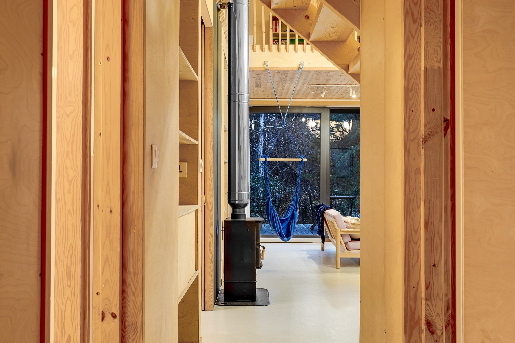 Wnętrze domu z drewnianym wykończeniem, piecem opalanym drewnem i wiszącym hamakiem z widokiem na las przez okno.