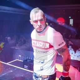 Szymon Citak - Zawodnik MMA z tatuażami na ramieniu, w białej koszulce z napisem OCTAGON, na ringu przed publicznością.