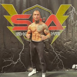 Szymon Citak - Mężczyzna z tatuażami na torsie i szyi, pozujący na tle logotypu sali sportów walki w sportowych spodniach i skarpetkach.