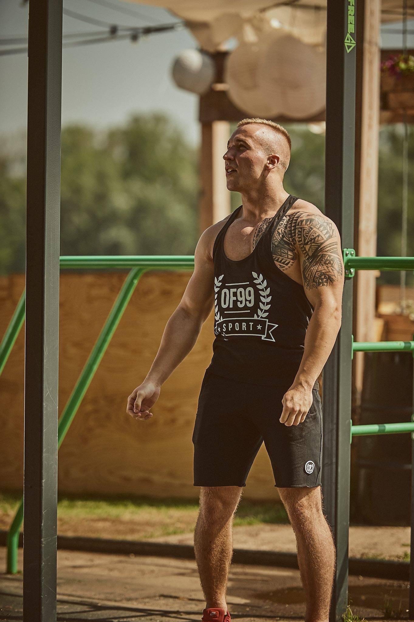 Mężczyzna z tatuażem na ramieniu w czarnym sportowym stroju stoi obok konstrukcji do ćwiczeń na świeżym powietrzu, spoglądając w lewo.