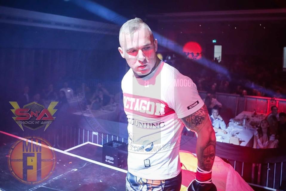 Zawodnik MMA z tatuażami na ramieniu, w białej koszulce z napisem OCTAGON, na ringu przed publicznością.