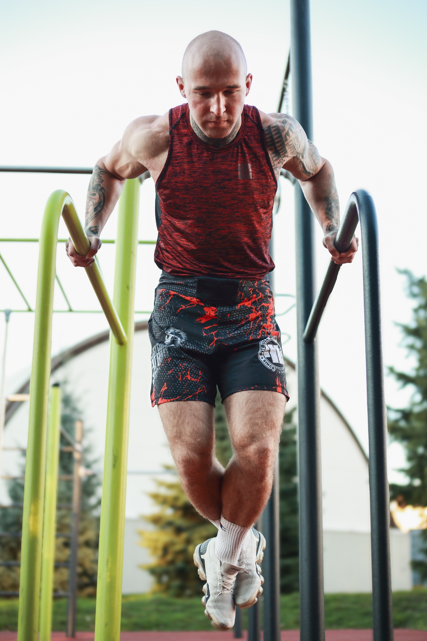 Mężczyzna z tatuażami na rękach wykonuje ćwiczenie na poręczach na zewnątrz, ubrany w sportowy strój.