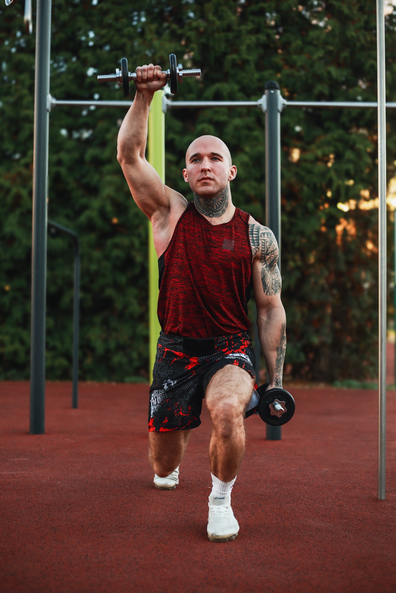 Łysy mężczyzna z tatuażami na szyi i ramionach wykonuje wykrok z hantlami na zewnątrz, na czerwonej nawierzchni placu do ćwiczeń, w tle zielone drzewa i drabinki.