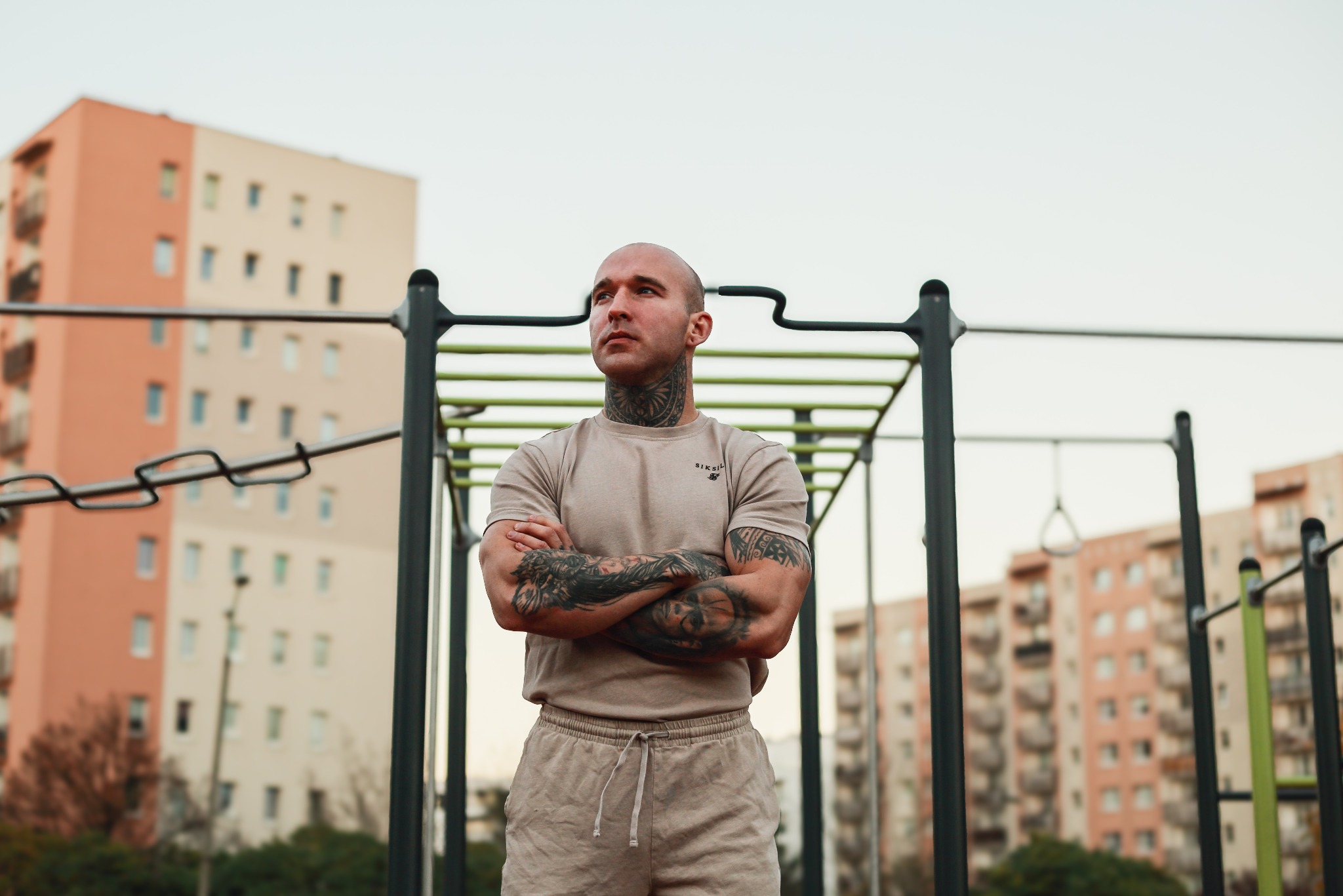 Mężczyzna z tatuażami na szyi i ramionach, ubrany w dres, stoi na tle drabinek do ćwiczeń na świeżym powietrzu, z budynkami mieszkalnymi w tle.