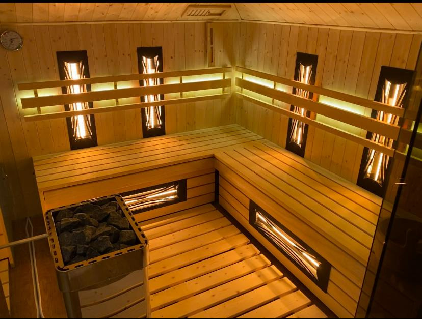 Wnętrze sauny wyłożone jasnym drewnem, z podświetlanymi panelami dekoracyjnymi i piecem wypełnionym kamieniami w metalowej obudowie.
