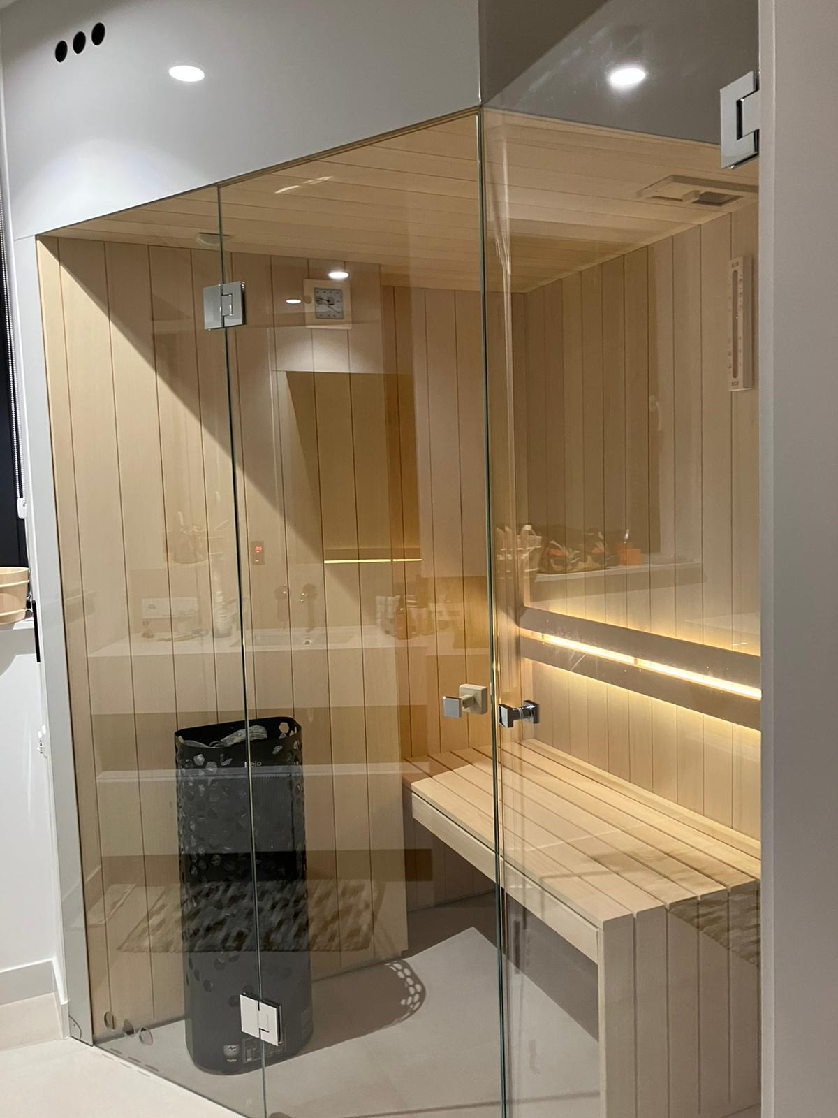 Nowoczesna kabina sauny z przeszklonymi ścianami, widoczny piec, drewniane ławki z podświetleniem LED oraz umywalka w tle.