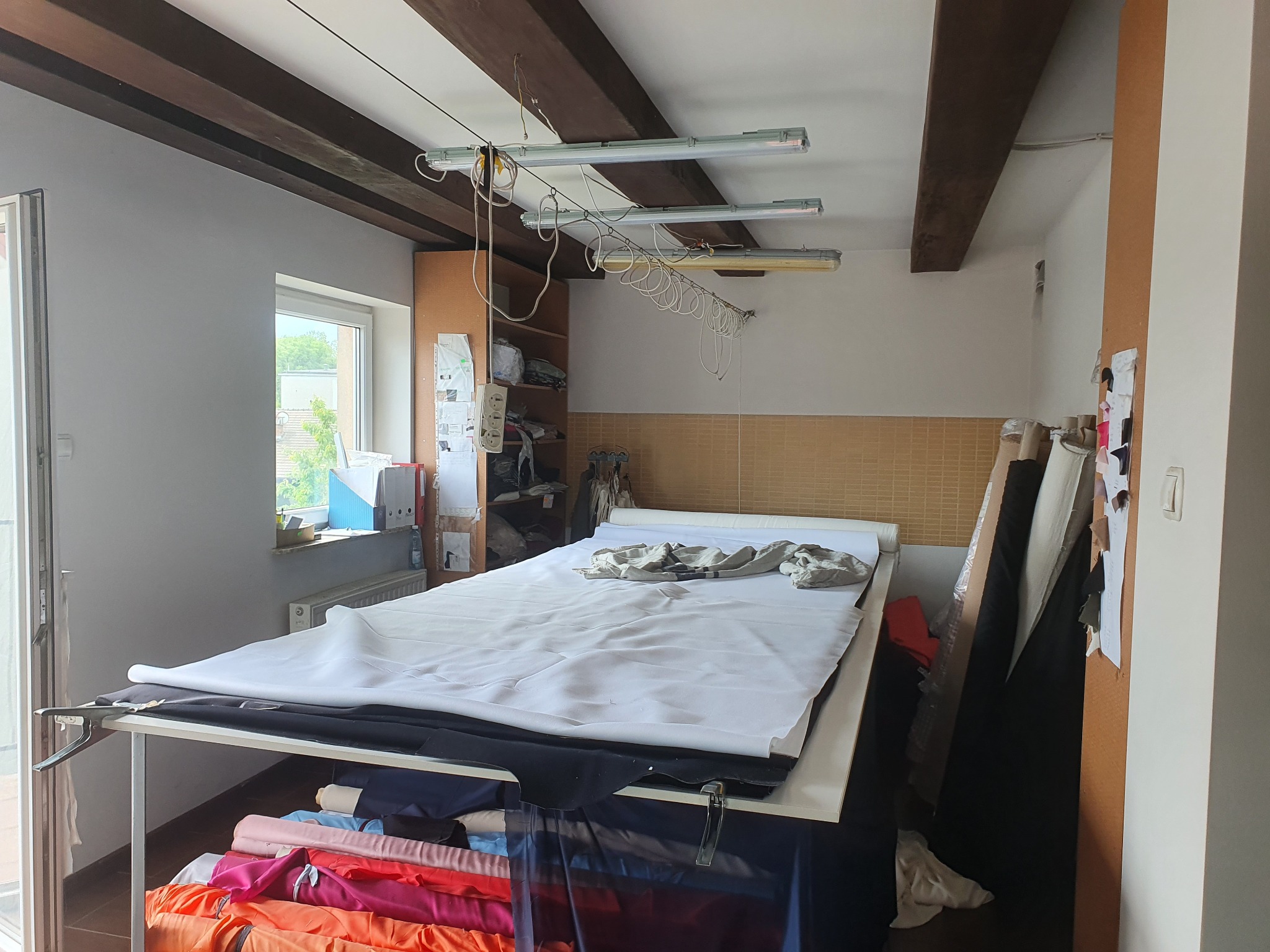 Duży stół krojczy z rozłożonym białym materiałem, pod spodem widoczne kolorowe bele tkanin, w tle regał z materiałami i lampy sufitowe w pracowni.
