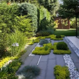 Nowoczesny ogród z szarymi płytami chodnikowymi, obłożony drobnymi kamieniami i ozdobnymi krzewami oraz trawami, z widoczną architekturą budynku w tle.