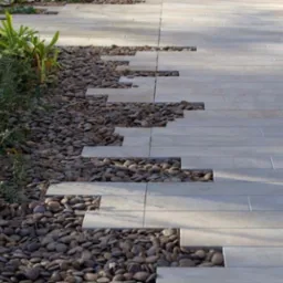 Ułożona ścieżka z jasnych, prostokątnych płyt chodnikowych z nieregularnymi przerwami wypełnionymi ciemnymi, okrągłymi kamieniami, obok rosną zielone rośliny.