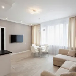 Jasne wnętrze salonu z beżową sofą narożną, okrągłym białym stołem z czterema krzesłami, telewizorem na ścianie i zasłonami w odcieniach beżu.