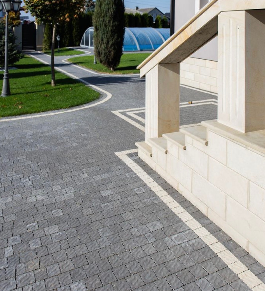 Szara kostka brukowa ułożona we wzór jodełki, obramowana jasną kostką, prowadząca do wejścia z jasnymi kamiennymi schodami i kolumnami, w tle trawnik i zadaszony basen.