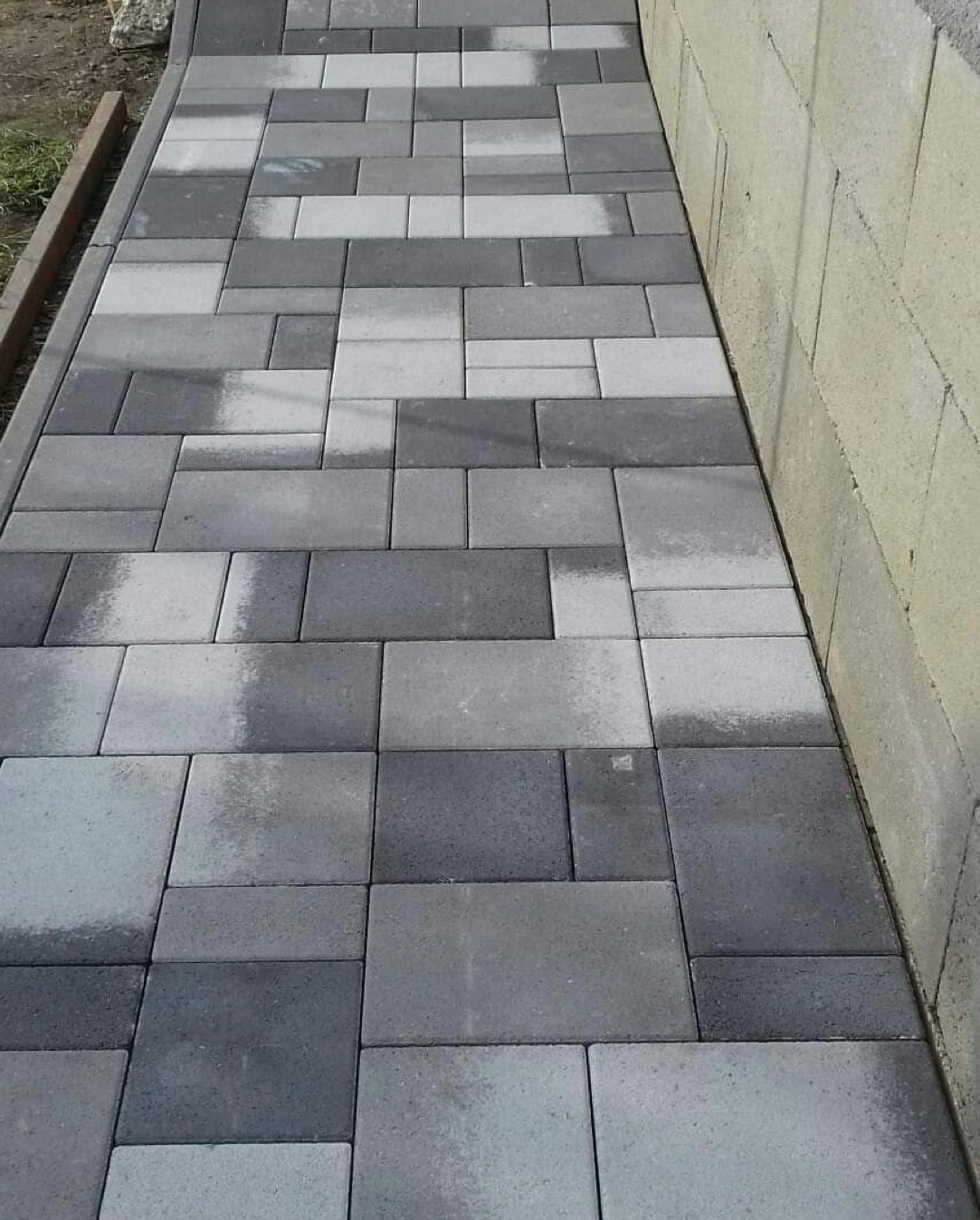 Świeżo ułożony chodnik z kostki betonowej w odcieniach szarości, przylegający do ściany z bloczków betonowych. Widoczne różne rozmiary i odcienie kostek tworzące wzór. Krawężnik oddziela chodnik...