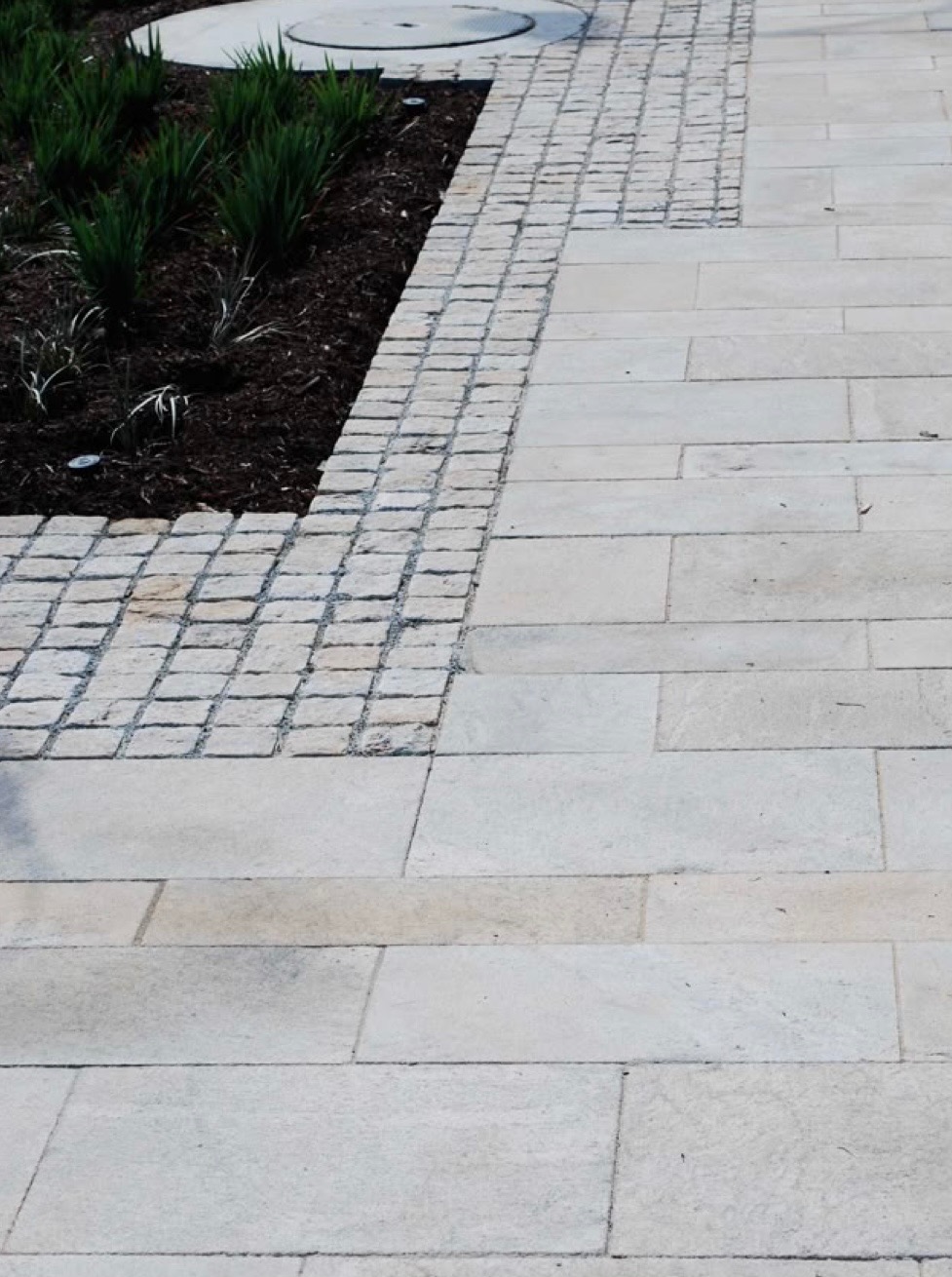 Jasnoszara nawierzchnia z prostokątnych płyt, obrzeża z kostki granitowej i fragment rabaty z zielonymi roślinami, widoczny właz kanalizacyjny w tle.