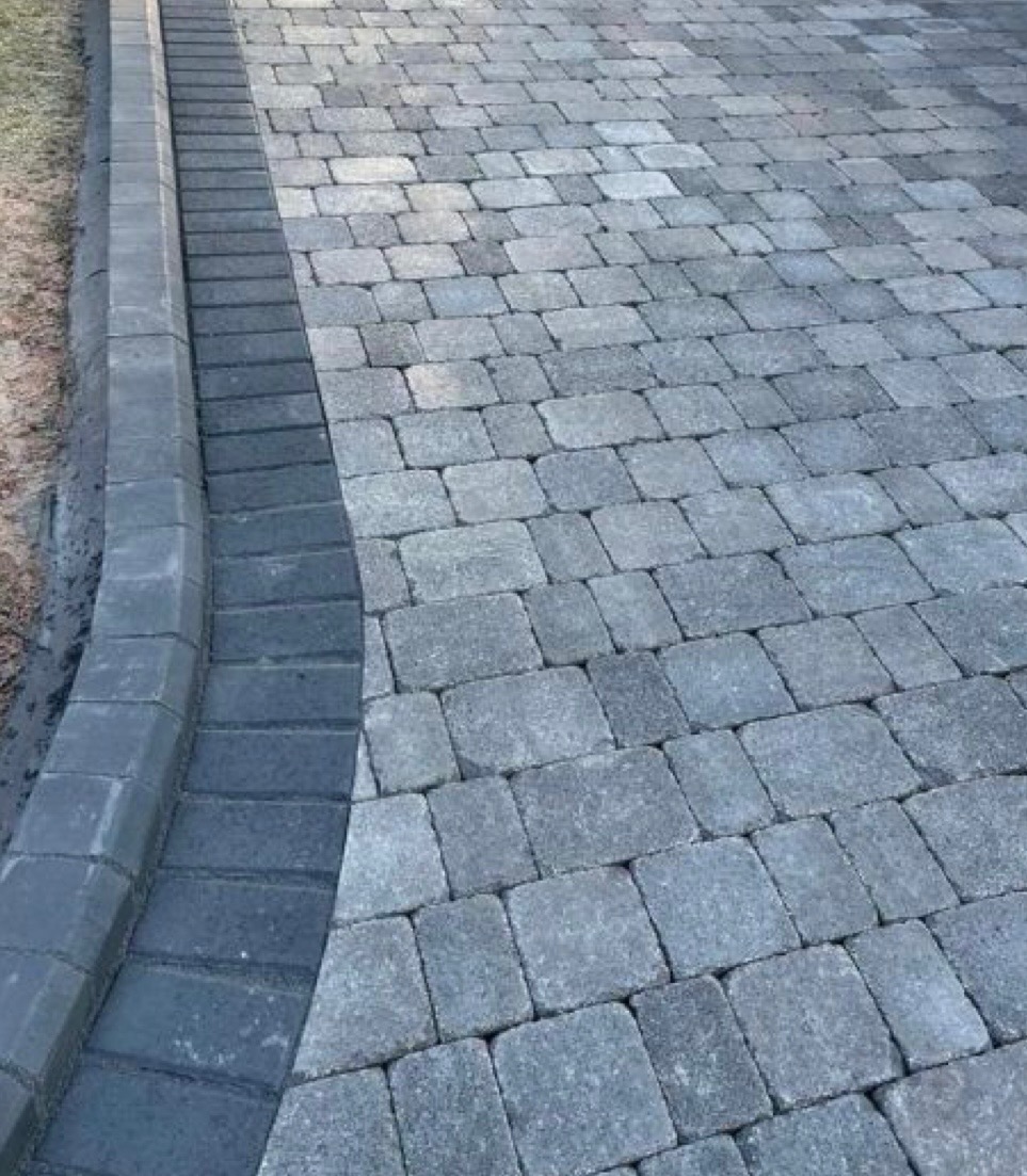 Świeżo ułożona, szara kostka brukowa z obrzeżem z ciemniejszych kostek, widoczna delikatna linia oddzielająca kostkę od gruntu.